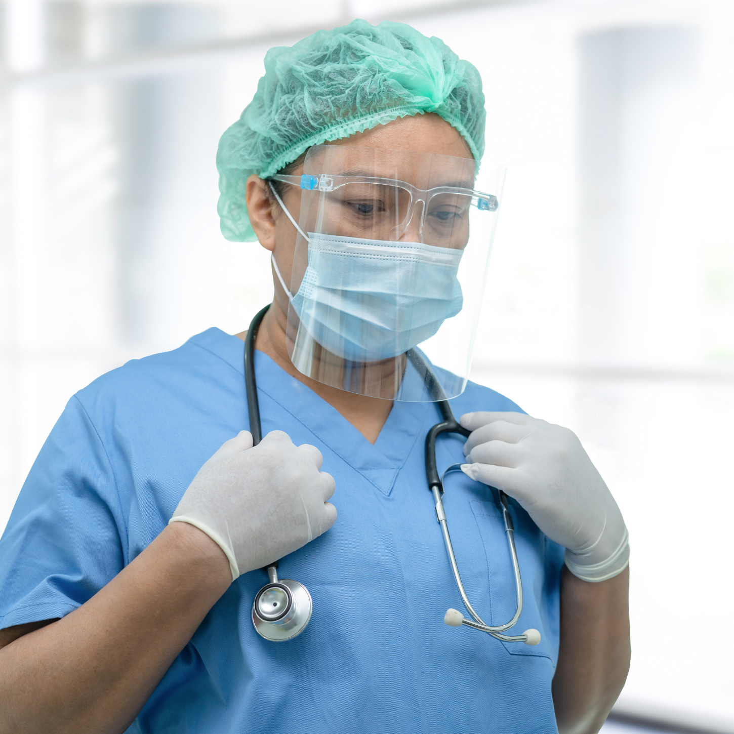 Nurse in blue scrubs, surgical mask, gloves, disposable green hair cap, clear face shield
